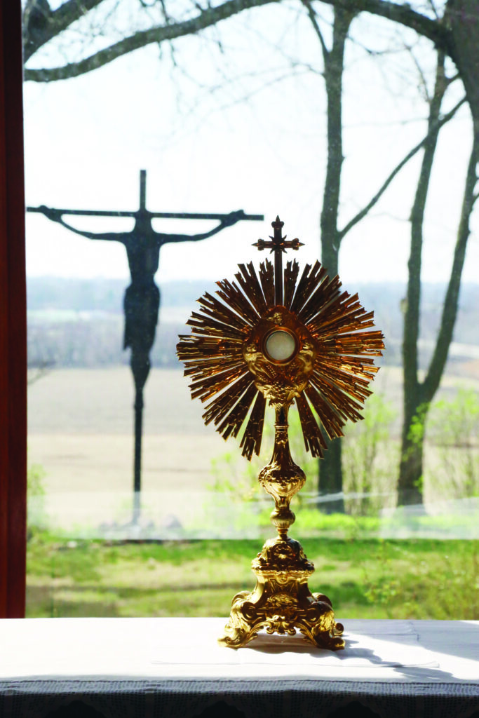 Eucharistic adoration monstrance at Mir House of Prayer in St. Joseph, with crucifix silhouette in the background seen through the window outdoors behind the monstrance.