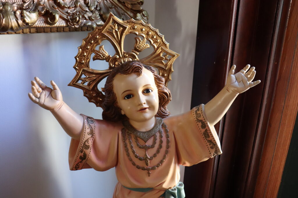 Baby Jesus statue at Alexandra's House: an infant Jesus reaches out to be picked up, dressed in a pink robe with a golden cruciform halo.